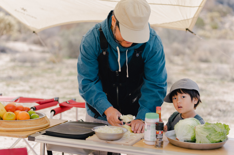 A father demonstrates how to cook on his IGT Camp Kitchen while his small child looks on.