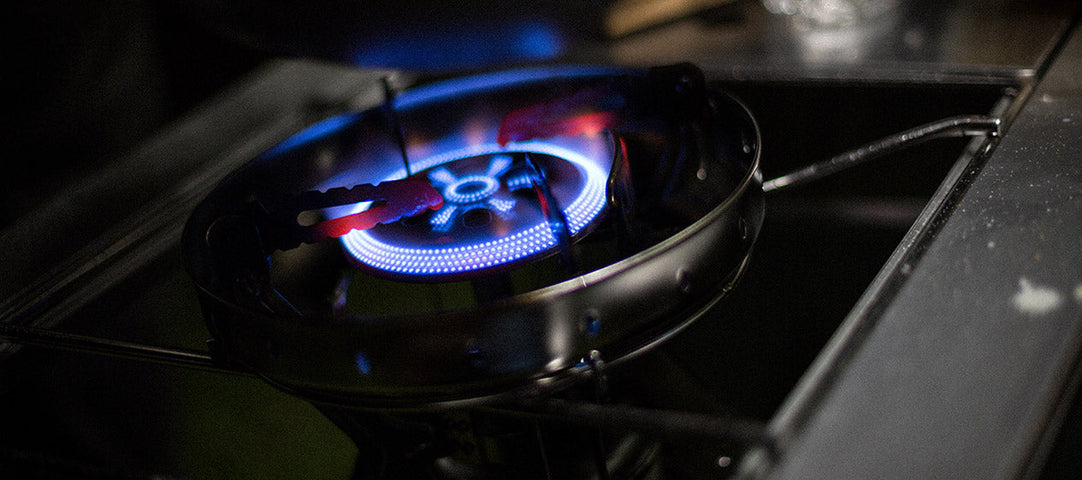 Close-up of a gas burner with blue flame on a camping stove at night.