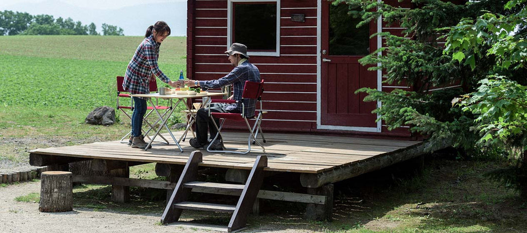 Two people setting a table on a wooden deck outside a small red cabin in a rural area.