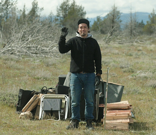 A Snow Peak gear expert stands in front of camping equipment in a field smiling and waving.