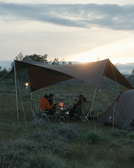 A group sits around a campfire under a Snow Peak tarp at sunset.