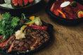 A cast iron pan is filled with grilled meat and vegetables. Two other cast iron pans with cooked food sit in the background.