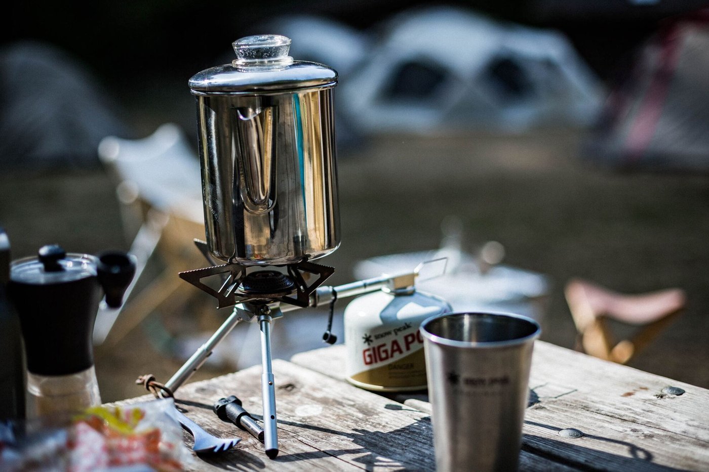 A camping stove is being used outdoors to heat a coffee pot. A camping mug and a canister of fuel are also visible on the table.