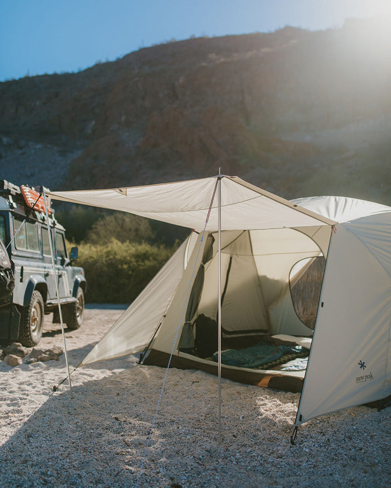 A white tent with a connected tarp is set up in a desert landscape. The tarp provides additional shade and cover for the tent. A vehicle is parked nearby.